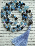 Full view of mala with 8mm/6mm translucent Porcelain Blue Agate; faceted 8mm/6mm light Blue Jade; 8mm blue Mosaic Quartz (dark blue with Bronzite inclusions); 6mm Rainbow Quartz Crystal beads and 16mm spiky silver "disco ball" guru with pale blue sutra and tassel. This one-of-a-kind mala is hand-knotted.