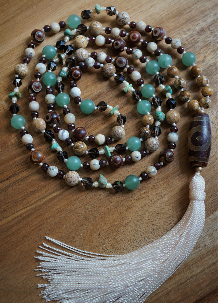Full view of mala with 8mm and 6mm Picture Jasper (light brown with dark brown speckles, spots, and lines); 8mm light green Adventurine; translucent brown 6mm faceted Smoky Quartz; 6mm White African Opal framed with deep red 4mm Garnet beads; Turquoise chip beads framed with ivory seed beads; 8mm red and light brown Dzi Agate rounds and 10x30mm barrel-shaped guru. A vanilla cream sutra and tassel bring this eclectic design full circle.