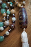 Close up view of 10x30mm barrel-shaped Dzi Agate guru. Deep red with light brown accents. A vanilla cream tassel spills from the base of this bead.