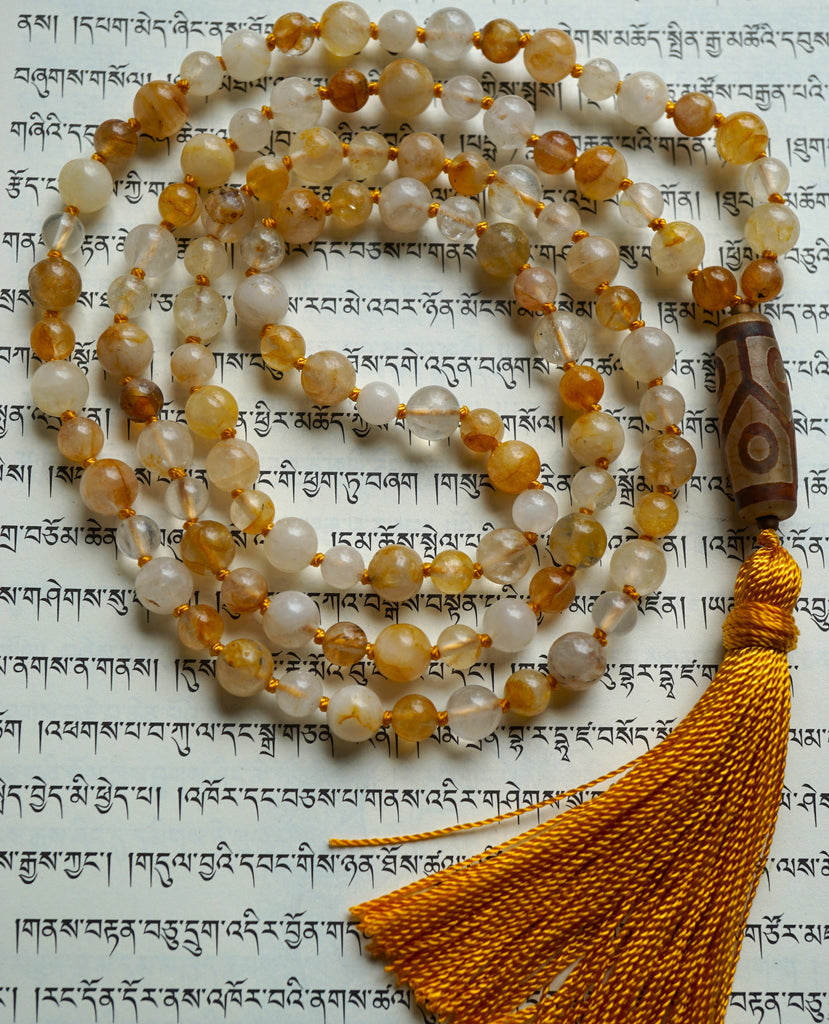 Full view of Golden Healer Quartz Mala. Alternating 8mm and 6mm Golden Healer Quartz beads. Range in color from white, pale yellow, soft gold, to brown. A 10x24mm Dzi Agate guru with light and dark brown colors and a geometric design brings this design full circle. A honey gold tassel spills from the base of this hand-knotted design.