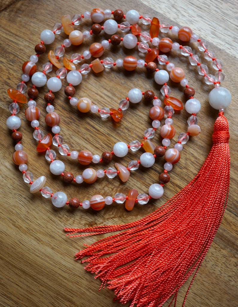 Full view of mala. White Moonstone; clear, star-cut Quartz Crystals; 8x13 orange Chalcedony chip beads; 6mm brownish orange Goldstone beads with sparkly copper flecks; 8mm Orange Agate beads framed with 4mm white Moonstone. This hand-knotted mala includes a terra cotta sutra and tassel that spills from the base of the 14mm Moonstone guru.