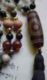 Close up view of 12x32mm barrel-shaped Dzi Agate guru (brown and red). Spilling from the base of the guru is a black tassel.