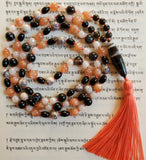 Full view of Peachy Keen Mala. 8mm round Peach Moonstone beads, 8mm dark black Sardonyx beads with white lines and whorls. 6x8mm rice-shaped Onyx beads add visual interest. 8mm Porcelain Jasper with cream, gray, and black inclusions. 6mm faceted White Jade, 6mm Golden Obsidian beads near the base of the mala, and 8x40mm oblong Sardonyx guru, black with white lines and inclusions. A beautiful peach sutra (cord) runs through all of the beads, and a peach tassel spills from the base of the guru.