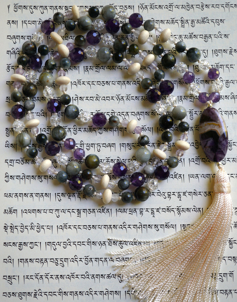 Full view of mala with dark purple Amethyst, light purple Lavender Amethyst, gray Cat's Eye, white Crackle Quartz, and cream-colored Pine rondelles. An oval 15x30mm Chevron Amethyst guru brings this design full circle with a cream-colored tassel spilling from the base of the bead.