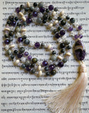 Full view of mala with dark purple Amethyst, light purple Lavender Amethyst, gray Cat's Eye, white Crackle Quartz, and cream-colored Pine rondelles. An oval 15x30mm Chevron Amethyst guru brings this design full circle with a cream-colored tassel spilling from the base of the bead.