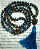 Full view of mala; 8mm blue and brown mosaic quartz with dark brown Bronzite beads; 6mm faceted Blue Agate beads mark the quarter and three-quarter mark for visual interest. An oval-shaped Eagle Eye guru with brown and gray striations brings this handsome mala full circle with a marine blue tassel spilling from the base.