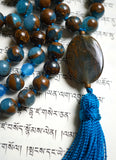 Close up view of oval Eagle Eye guru in soft gray and dark brown, which complements the Blue Mosaic Quartz and Bronzite beads of this mala design. A marine blue tassel spills from the base of this guru bead.