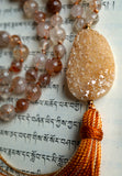 Close up view of cream-colored Druzy Agate guru. The tiny quartz crystals (druzy) symbolize inner strength, renewal, and clarity. They also capture and reflect light. A variegated cinnamon brown tassel spills from the base of this focal bead.