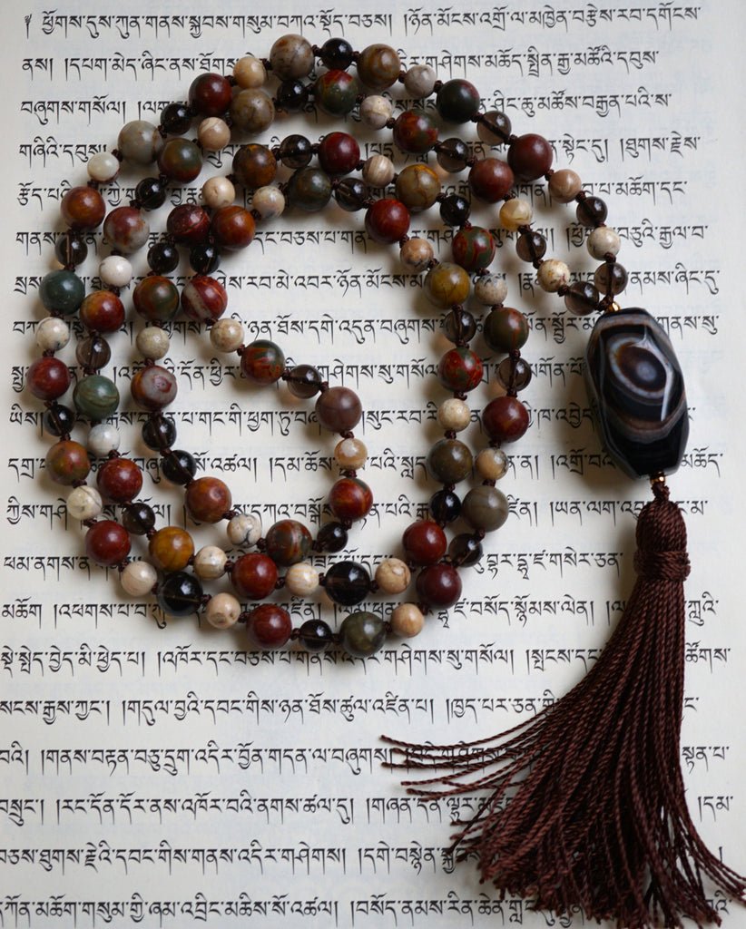 Full view of mala with transparent brown Smoky Quartz; red, gray, and brown Red Creek Jasper; cream-colored White African Opal beads; a faceted barrel-shaped Brown Agate guru and chocolate brown sutra and tassel.
