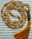 Full view of Golden Healer Quartz Mala. Alternating 8mm and 6mm Golden Healer Quartz beads. Range in color from white, pale yellow, soft gold, to brown. A 10x24mm Dzi Agate guru with light and dark brown colors and a geometric design brings this design full circle. A honey gold tassel spills from the base of this hand-knotted design.