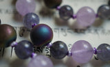 Close up view of 8mm Druzy Agate beads, which include small crystal structures that form on the stone. These beads are dark purple with a gold and dark blue sheen.  They combine beautifully with the violet-gray Iolite and light purple Lavender Amethyst beads in this mala design.