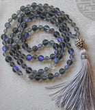 Full view of mala. Mystic Aura beads are treated Quartz beads that create a flash of color on their surface. These soft grey beads with iridescent blue flashing catch  light and attention. Two 8mm carved Crystal Quartz beads mark the quarter and three-quarter mark. A silver "disco ball" guru brings this design full circle along with a dove gray tassel.