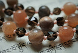 Close up view of Peach Moonstone beads. Some beads are a solid peach color with pinkish-peach flecks. Some are light peach with gray. Both varieties come together with the brown Smoky Quartz beads .  The light peach sutra connects all of the beads together. The knots between protect and showcase the beads in this design.