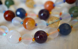 Close up view of 8mm purple Amethyst, white Moonstone, faceted Blue Agate, orange Agate, faceted yellow Citrine, green African Jade, and red Apple Jasper beads; 6mm Mystic Aura Crystal beads frame the 8mm beads of this design. Variegated rainbow knots protect and showcase the beads of this design beautifully.