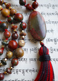 Close up view of 15x30mm oval Red Creek Jasper guru with red, gray, and brown hues. It brings this design full circle, with a variegated red and black tassel spilling from the base.