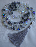 Full view of Simply Elegant Mala. 6mm and 8mm Mystic Aura beads (clear, matte white, clear blue, and matte gray). These glass beads have been treated so that an indigo flashing from the core catches with light. A simple metal guru brings this design full circle, along with a dove gray tassel.