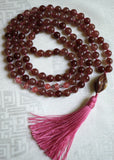 Full view of mala with purplish, burgundy red Strawberry Quartz beads. Pink Cherry Quartz beads mark the quarter and three-quarter marks of the mala. A twisted spiral Mookaite Jasper guru brings this hand-knotted design full circle. A dusty rose tassel spills from the base of the guru.
