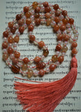Full view of Summer Solstice Mala. 8mm Red Botswana Agate beads range in color from red, orange, and peach. These beads with white bands complement the 6mm faceted White Agate beads. Two 10x14mm Red Carnelian beads framed with clear Swarovski crystals mark the quarter and three-quarter mark of this design. Two 8mm Red Botswana Agate beads function as the guru. A soft salmon colored sutra and tassel bring this beautiful design full circle.