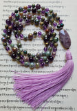 Full view of purple Amethyst and faceted, rainbow-plated Jasper beads that shimmer in shades of light green, lavender, and light brown. A purple and white Chevron Amethyst guru with lavender sutra and tassel brings this design full circle.