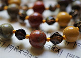Close up view of 8mm Apple Jasper (red with gray and brown inclusions), faceted 6mm Smoky Quartz, and 8mm Golden Mookaite beads. Each bead is separated with a knot to protect  and showcase the beads.