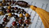 Close up view of beads, tassel, and guru. Goldenrod tassel is hand made and directly connected to the sutra that binds all the beads together. 8mm Apple Jasper (red with brown and gray inclusions), Golden Mookaite; 6mm Apple Jasper and faceted translucent brown Smoky Quartz beads with barrel-shaped Dzi Agate guru (rustic red, brown, and gray). Warm, earthy hues just in time for fall.