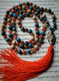 Full view of hand-knotted Fire and Sky Mala with brilliant orange Fire Agate, bright blue Apatite, turquoise green Cuprite, and earthy, natural tones of Fossil Jasper. A bold lampwork guru with bright orange and blue brings this design full circle with a fire orange sutra and tassel.