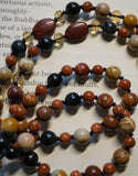Close up view of 8mm Mookaite Jasper beads; 6mm Goldstone and Blue Tiger Eye; two 10x14 red oval Mookaite Jasper beads framed with light gold crystals mark the quarter and three-quarter mark of the mala. Navy blue sutra (cord) and tassel. Hand-knotted mala.