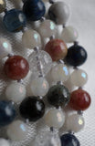 Vertical view of beads. 8mm Pink Tourmaline, Dark Blue Lapis Lazuli, white Howlite with soft gray inclusions, Black Tourmaline, Cracked Quartz Crystal, and light blue Kyanite. 6mm faceted White Jade beads separate and alternate among the larger beads. Hand-knotted mala.
