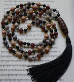 full view of Smoke and Fire Mala. 8mm red Strawberry Quartz, gray, pink, and black Porcelain Japer, and light brown Rosewood beads framed with 4mm faceted gold beads. 6mm black Golden Obsidian beads and gray faceted Labradorite beads combine with the barrel-shaped Dzi Agate guru (brown and red). A black sutra (cord) and tassel bring this hand-knotted design full circle. 