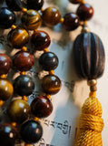 Close up view of dark brown barrel-shaped wood guru. Goldenrod tassel. Red, Blue, and Gold Tiger Eye beads.