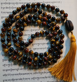full view of knotted mala. Red Tiger Eye. Gold Tiger Eye. Blue Tiger Eye. Goldenrod tassel. Carved dark brown wood guru.